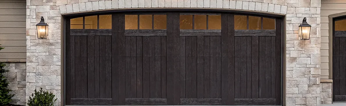 Dark brown, double garage doors with small window panels at the top, framed by light stone pillars and wall lanterns.